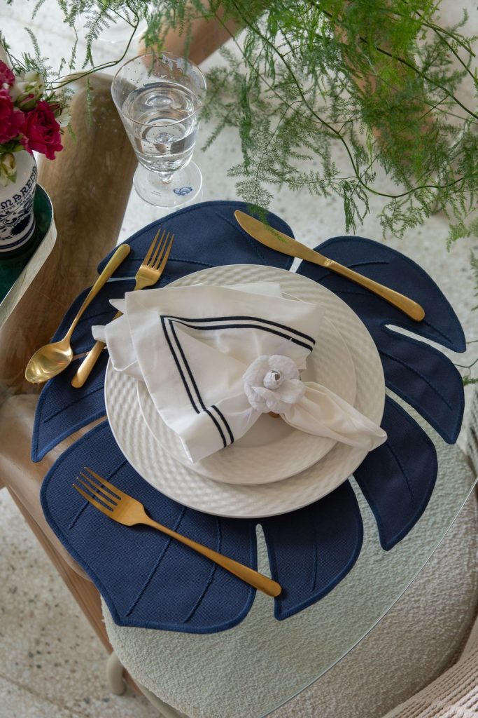 Mesa de vidro ao ar livre decorada com o duo de lugar americano e guardanapo marinho, em composição elegante e sofisticada.